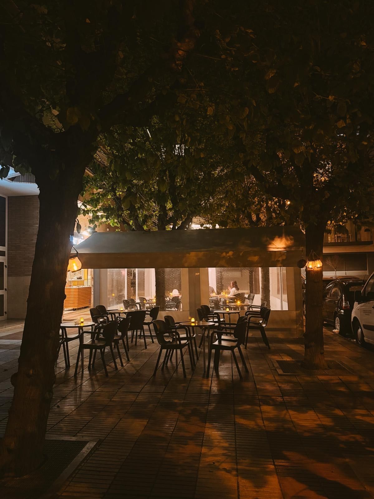 La terraza al anochecer, bajo los árboles, con velas en las mesas y el local iluminado al fondo